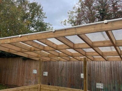 Freestanding timber shelter with polycarbonate roof and integrated bench seating in an EYFS playground