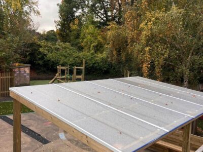 Aerial view of a freestanding timber shelter with polycarbonate roof and integrated bench seating in an EYFS playground